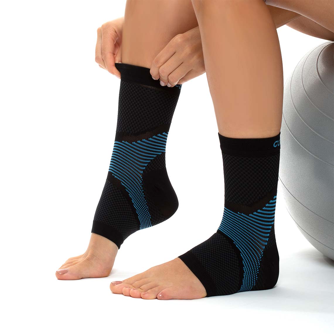 Nahaufnahme von zwei Beinen, auf denen eine Person schwarze, knöchelhohe CopperFit Ice Ankle Sleeve mit blauen Mustern überzieht; sie sitzt dabei neben einem Gymnastikball.