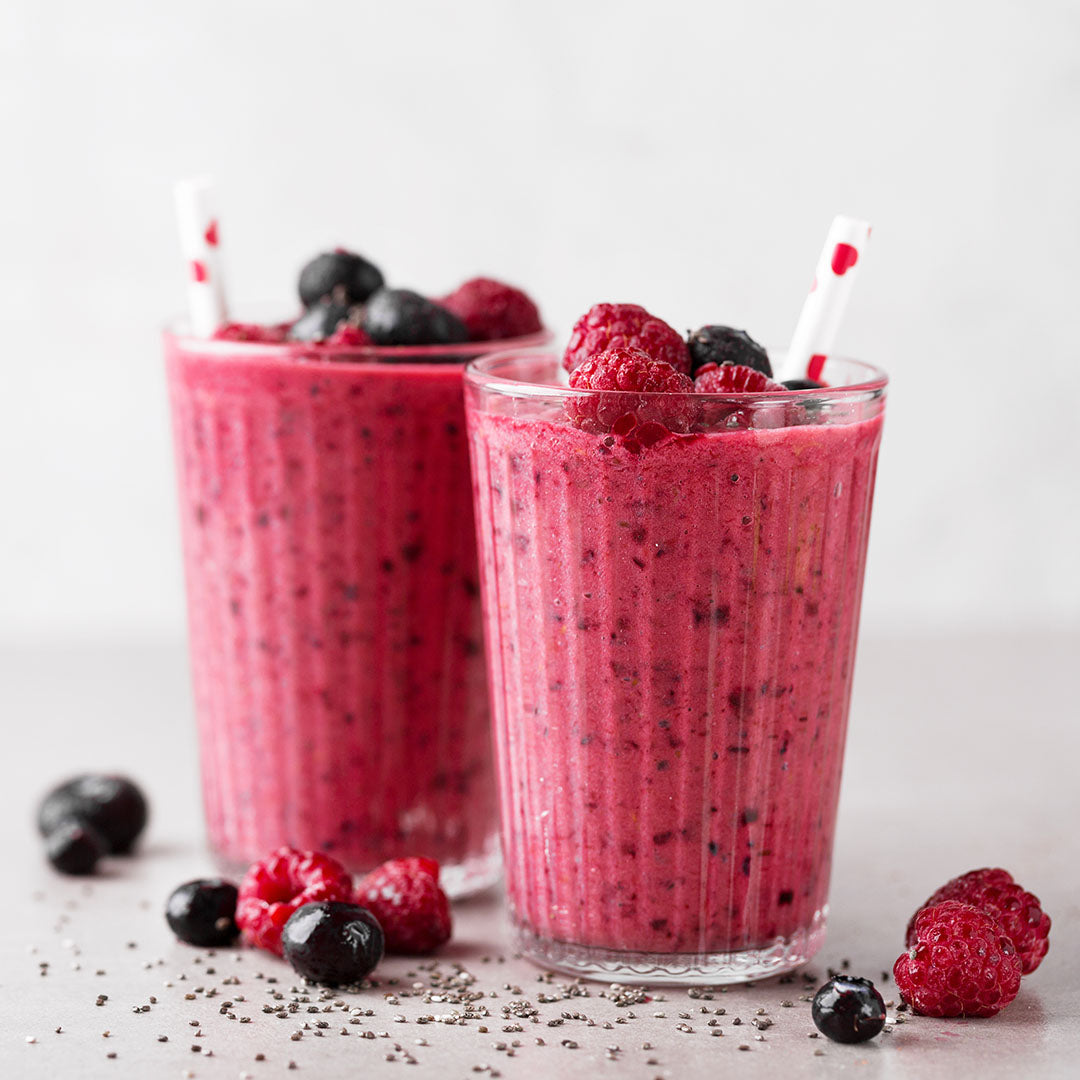Zwei Gläser mit cremigem Beeren-Smoothie, dekoriert mit Himbeeren und Heidelbeeren, daneben verstreute Chiasamen und Beeren.