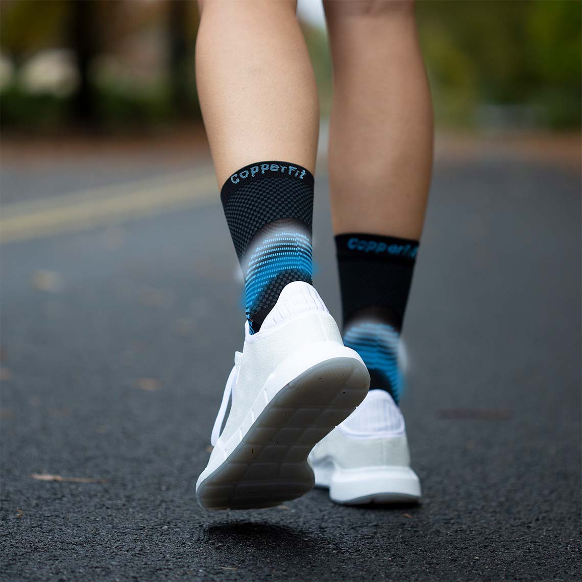 Rückansicht von Beinen einer laufenden Person auf einer Straße, getragen werden weiße Sportschuhe und schwarze Kompressionssocken mit blauem und grauem Farbverlauf sowie der Aufschrift „Copper Fit“.