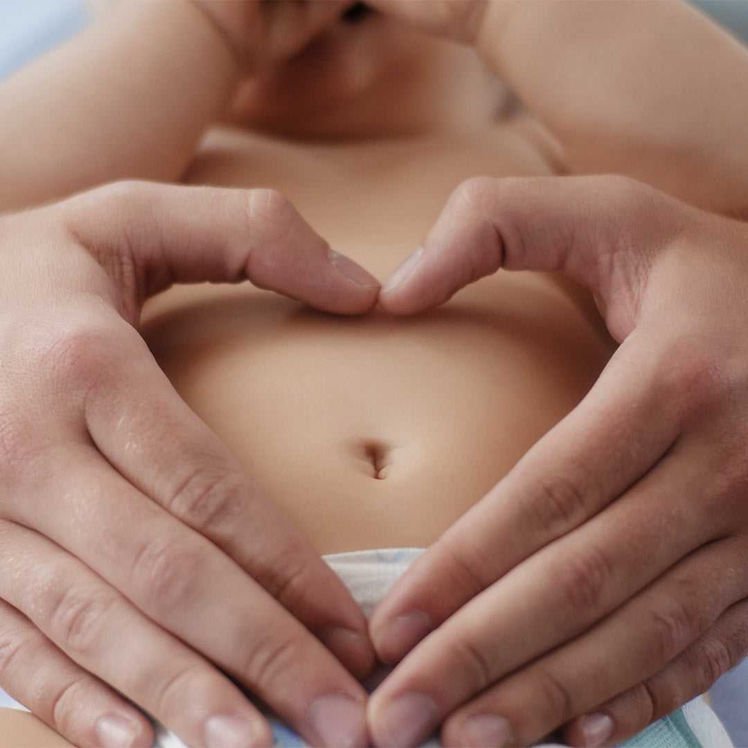 Zwei Hände formen liebevoll ein Herz um den Bauchnabel eines liegenden Babys – symbolisch für sanfte Nabelpflege und Babypflege.