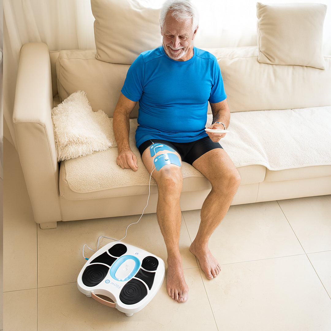 Älterer Mann sitzt auf einem Sofa, nutzt das Fllow Massagegerät mit Elektrodenpads am Bein und steuert es per Fernbedienung.