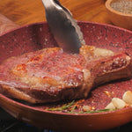 Ein Steak wird in der Copper and Stone Pan mit einer Zange angebraten, daneben liegen Knoblauch und Kräuter.