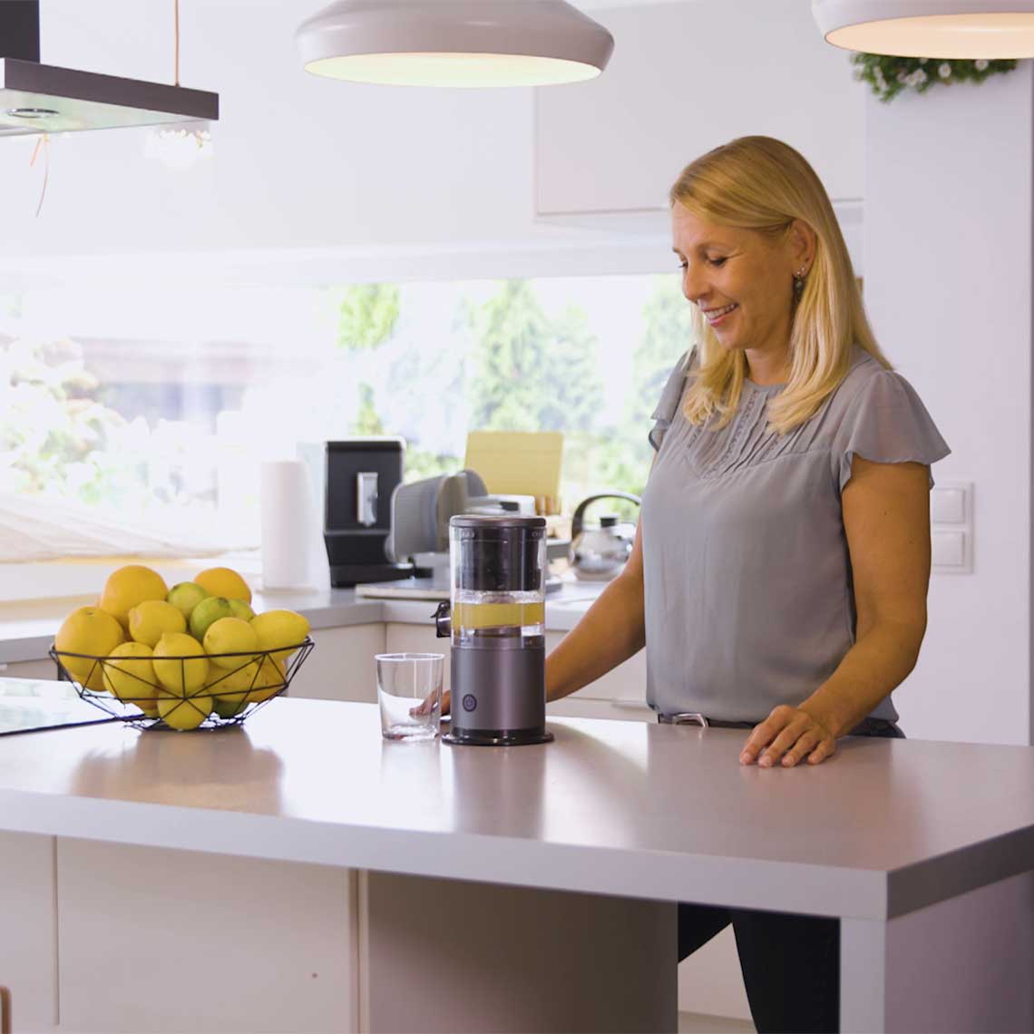 Eine Frau steht in der Küche und schaut auf den laufenden Livington JuiceBoost, daneben steht ein Glas mit Saft.