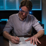 Ein Mann schreibt an einem Schreibtisch und trägt eine LED-Brille, die seinen Arbeitsbereich beleuchtet.