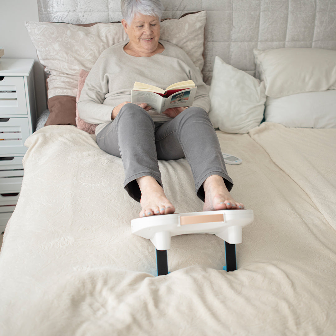 Ältere Frau liegt im Bett, liest ein Buch und nutzt dabei das Fllow Fußmassagegerät zur Entspannung.