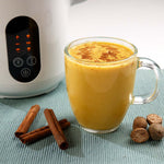 Glas mit goldener Gewürzmilch, daneben Zimtstangen und Muskatnüsse, im Hintergrund ein Pflanzendrink‑Maker.