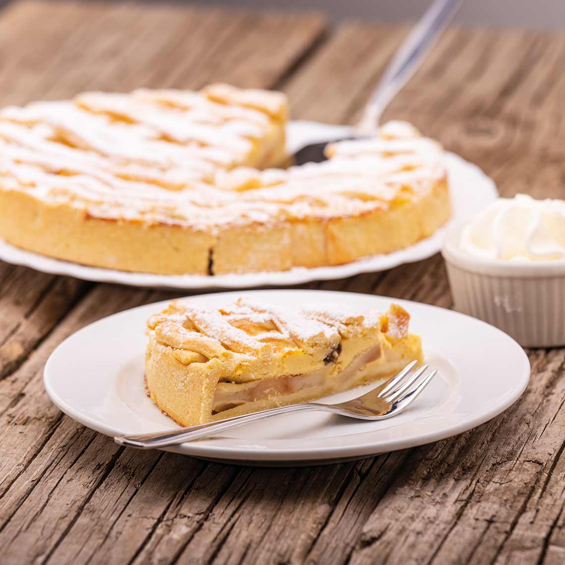 Ein Stück gedeckter Apfelkuchen mit Zimt und Puderzucker, serviert mit Schlagobers auf einem weißen Teller.