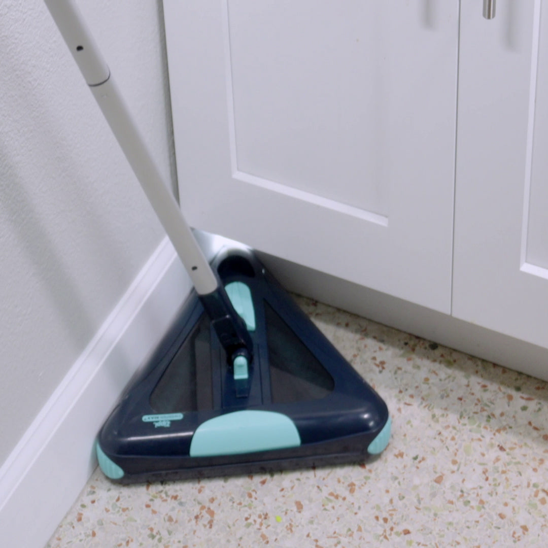 Ein dreieckiger E-Besen mit langem Stiel reinigt eine Ecke zwischen Wand und Küchenschrank auf einem hellen, gesprenkelten Boden.