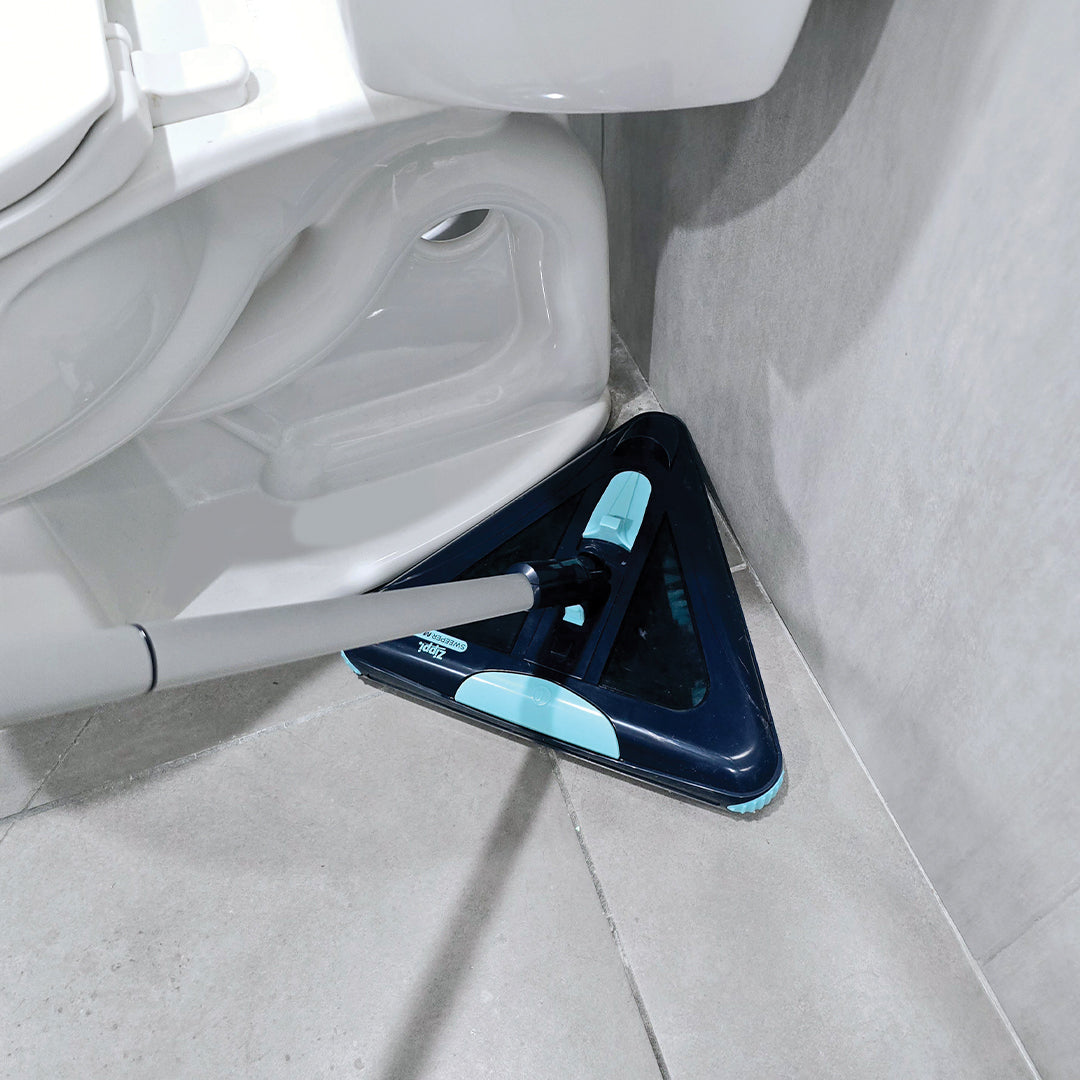 Ein dreieckiger Bodenwischer reinigt die enge Ecke neben einer Toilette auf hellen Fliesen und erreicht den Bereich zwischen Wand und Keramik.