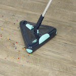 Ein dreieckiger E-Besen nimmt bunte Perlen von einem hellen Holzboden auf und zeigt die effektive Reinigung auch kleiner, verstreuter Verschmutzungen.