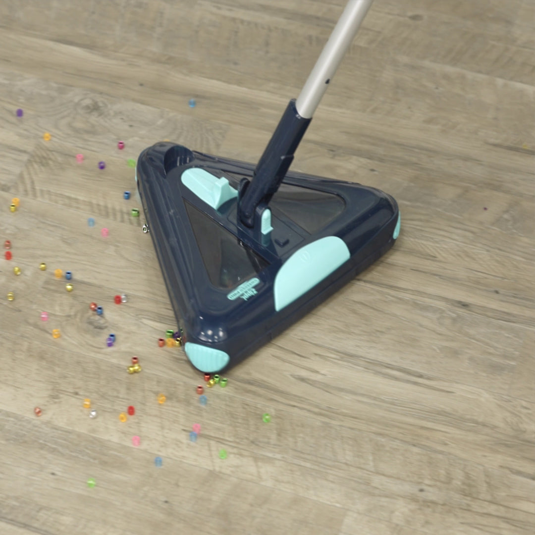 Ein dreieckiger E-Besen nimmt bunte Perlen von einem hellen Holzboden auf und zeigt die effektive Reinigung auch kleiner, verstreuter Verschmutzungen.