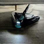 Ein dreieckiger Bodenreiniger fährt unter ein Möbelstück und beleuchtet den Boden dank integriertem LED-Licht, um Staub und Schmutz in dunklen, schwer erreichbaren Bereichen sichtbar zu machen.