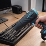 Eine Hand hält das kompakte AiroBlast Akku-Gebläse und bläst gezielt Staub von einer Computertastatur auf dem Schreibtisch.
