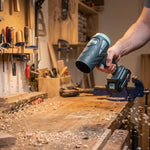 Eine Person bläst kraftvoll Holzspäne und Sägemehl von einer  Werkbank in der Werkstatt.