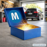Geöffnete blaue MediaShop Mystery Box Auto, aus der ein goldener Lichtschein strahlt, platziert auf einem Werkstattboden mit einem Fahrzeug im Hintergrund.