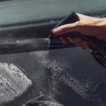 Eine Hand sprüht die wasserlose Platinum SpraySensation Autopflege mit einem elektrischen Sprühkopf auf eine dunkle Fahrzeugoberfläche. Feiner Sprühnebel bedeckt die Fläche, neben der ein Mikrofasertuch liegt.