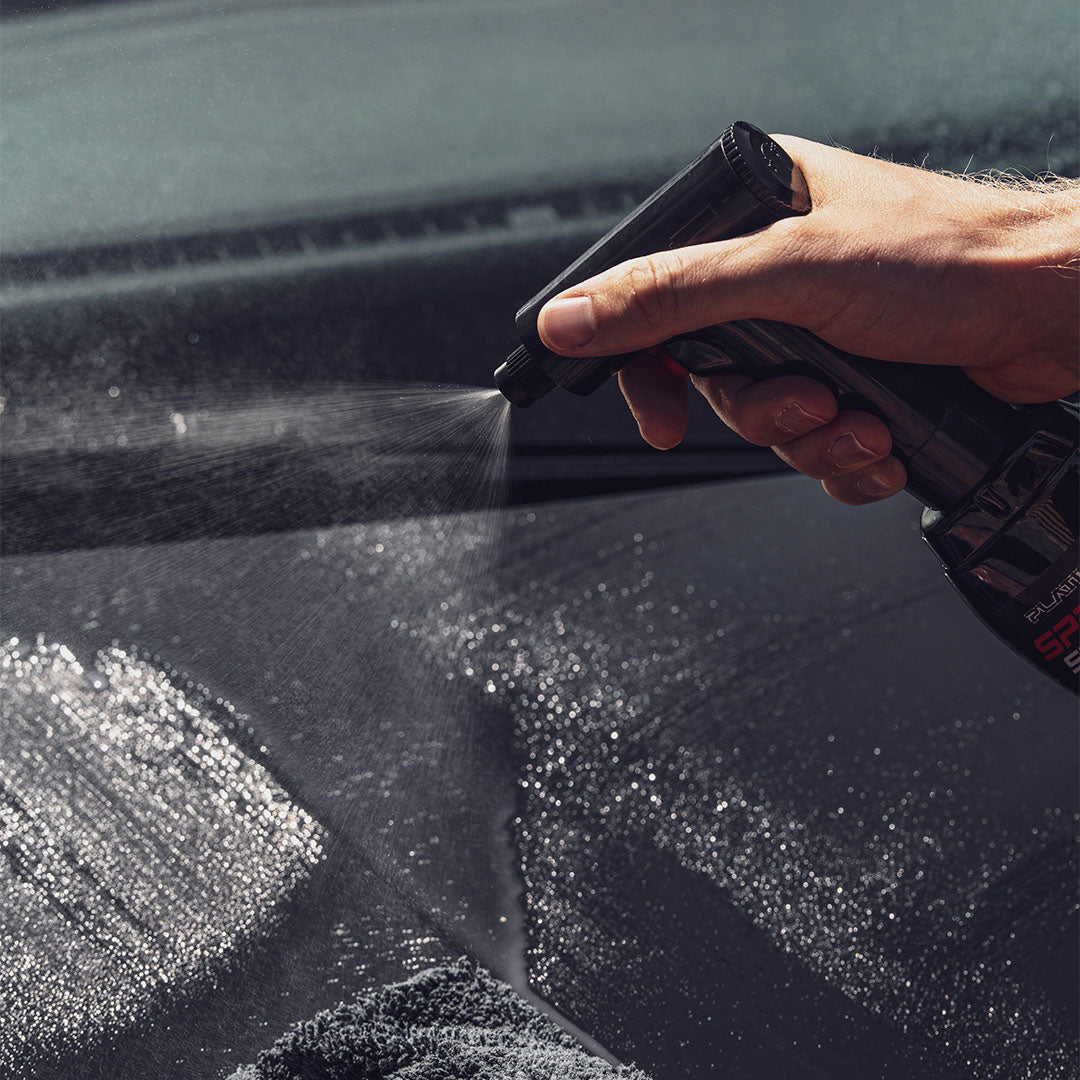 Eine Hand sprüht die wasserlose Platinum SpraySensation Autopflege mit einem elektrischen Sprühkopf auf eine dunkle Fahrzeugoberfläche. Feiner Sprühnebel bedeckt die Fläche, neben der ein Mikrofasertuch liegt.
