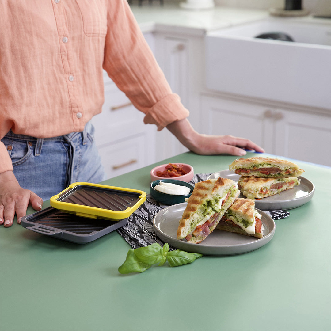 Eine Person steht in einer Küche neben dem Toastie Mikrowellen-Toaster. Auf Tellern liegen knusprige Paninis mit Pesto, Tomaten und Mozzarella, bereit zum Verzehr.