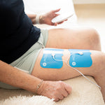 Person mit zwei blauen Elektrodenpads auf dem Oberschenkel, hält Fernbedienung zur Steuerung des Massagegeräts in der Hand.