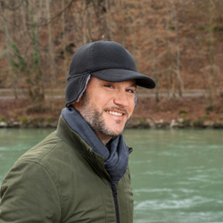 Mann mit Wintermütze und Schal steht lächelnd an einem Flussufer in herbstlicher Umgebung.