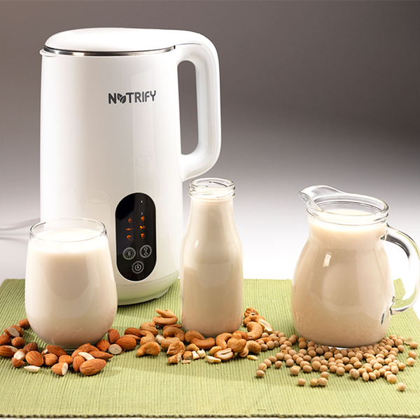 Pflanzendrink‑Maker mit Karaffen und Gläsern voller frisch zubereiteter Nussmilch, umgeben von Mandeln, Cashews und Sojabohnen.