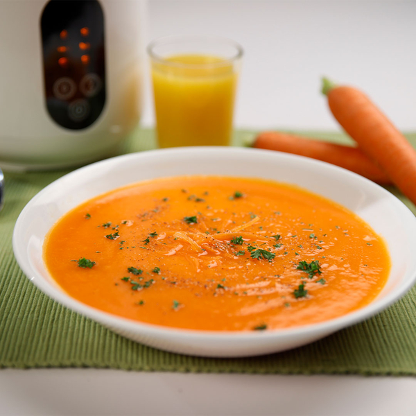 Schale mit cremiger Karottensuppe, garniert mit Kräutern, daneben frische Karotten und ein Glas Saft.