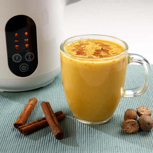 Glas mit goldener Gewürzmilch, daneben Zimtstangen und Muskatnüsse, im Hintergrund ein Pflanzendrink‑Maker.