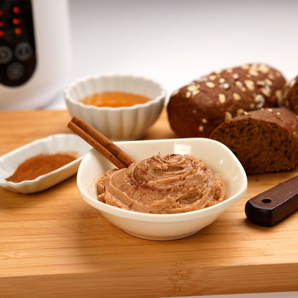 Auf einem Holzbrett steht eine Schale mit Aufstrich, dahinter eine Schale mit Zimt und ein dunkles Brot.