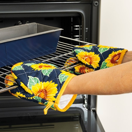 Person mit 2 Fäustlingen im Sonnenblumen-Design zieht damit einen Gitterrost aus dem Backofen